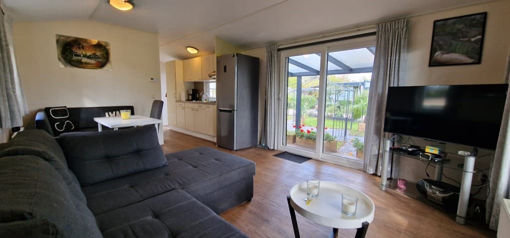 Gezellige woonkamer met een bank, tv, eettafel en toegang naar de tuin met een overkapping en planten.