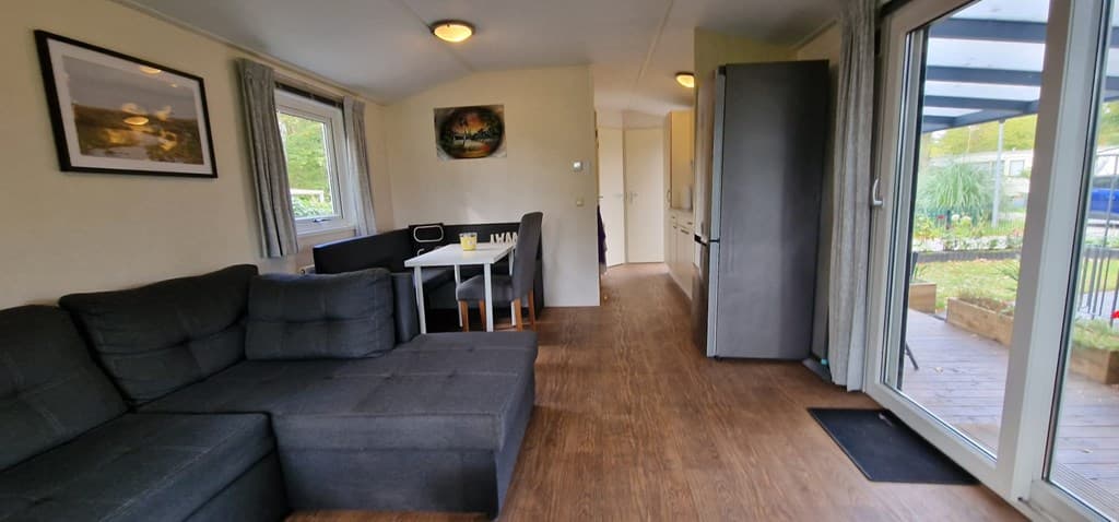 In de woonkamer staat een donkere hoekbank met een eettafel en stoelen, en er is een uitzicht op een terras buiten.