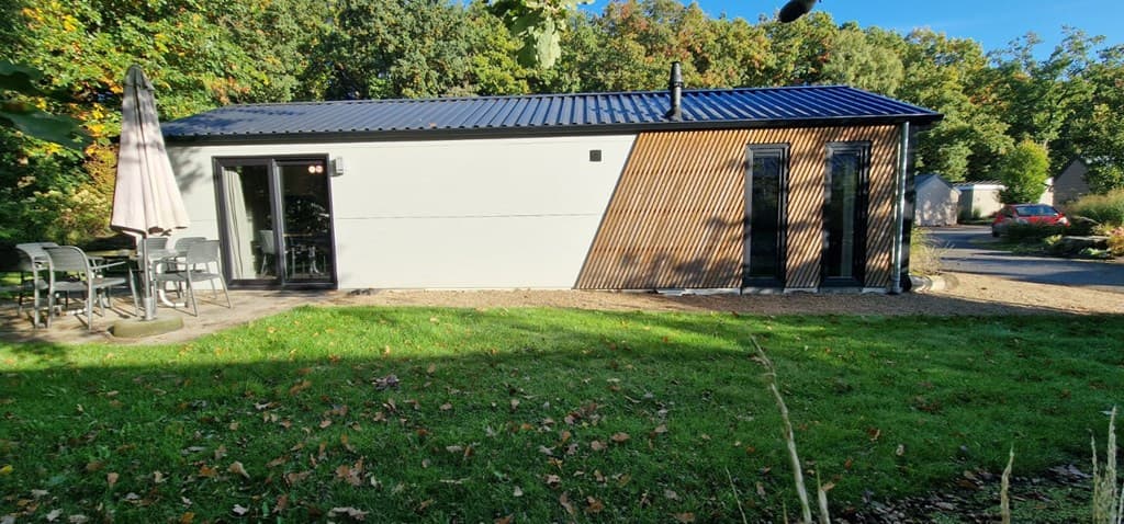 Moderne vakantiewoning met houten elementen, tuinmeubelen en een grasveld rondom in een bosrijke omgeving.