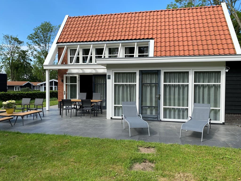 Vakantiewoning met een overdekt terras, moderne tuinstoelen en een grasveld voor de deur, omringd door bomen.