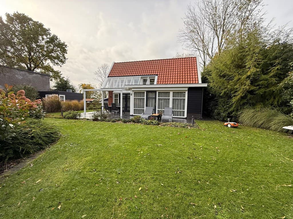Zwart houten vakantiehuis met rode dakpannen omringd door groene tuin en planten, terras met stoelen zichtbaar