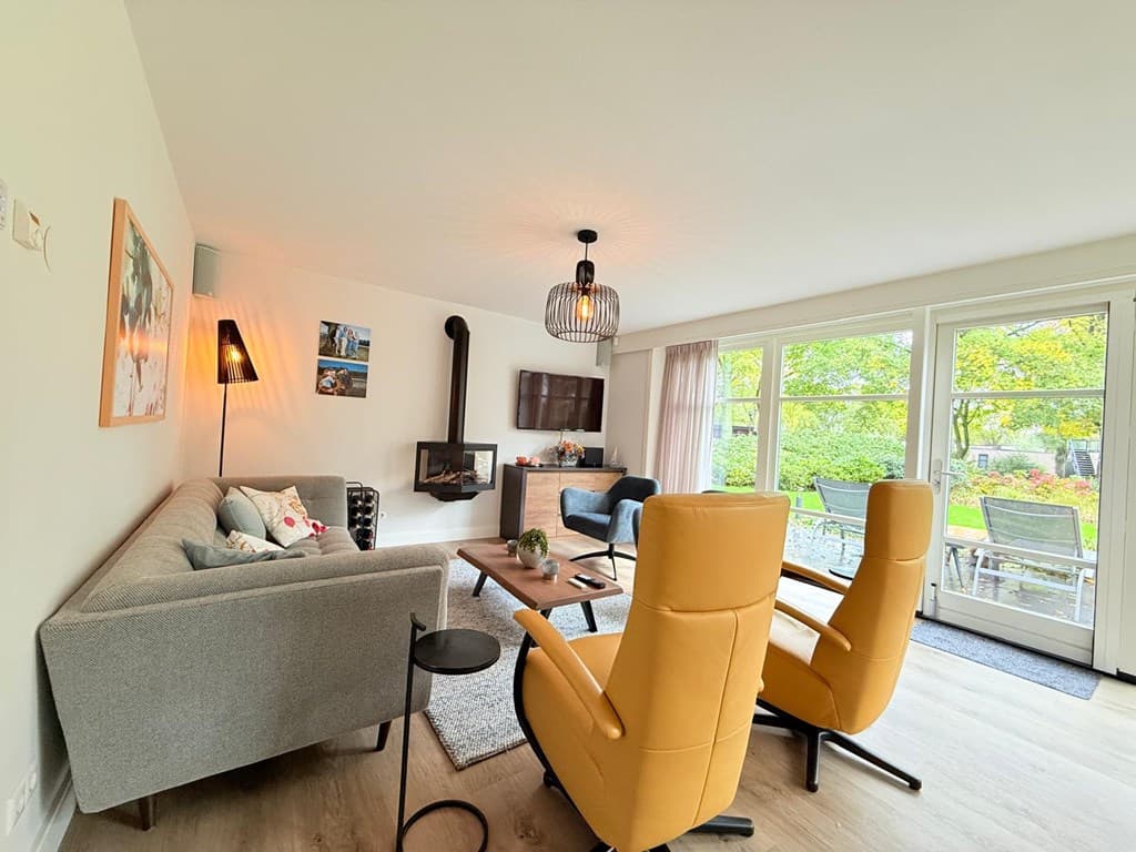 Lichte woonkamer met grijze en gele stoelen, een zithoek en uitzicht op de tuin door grote glazen schuifdeuren.