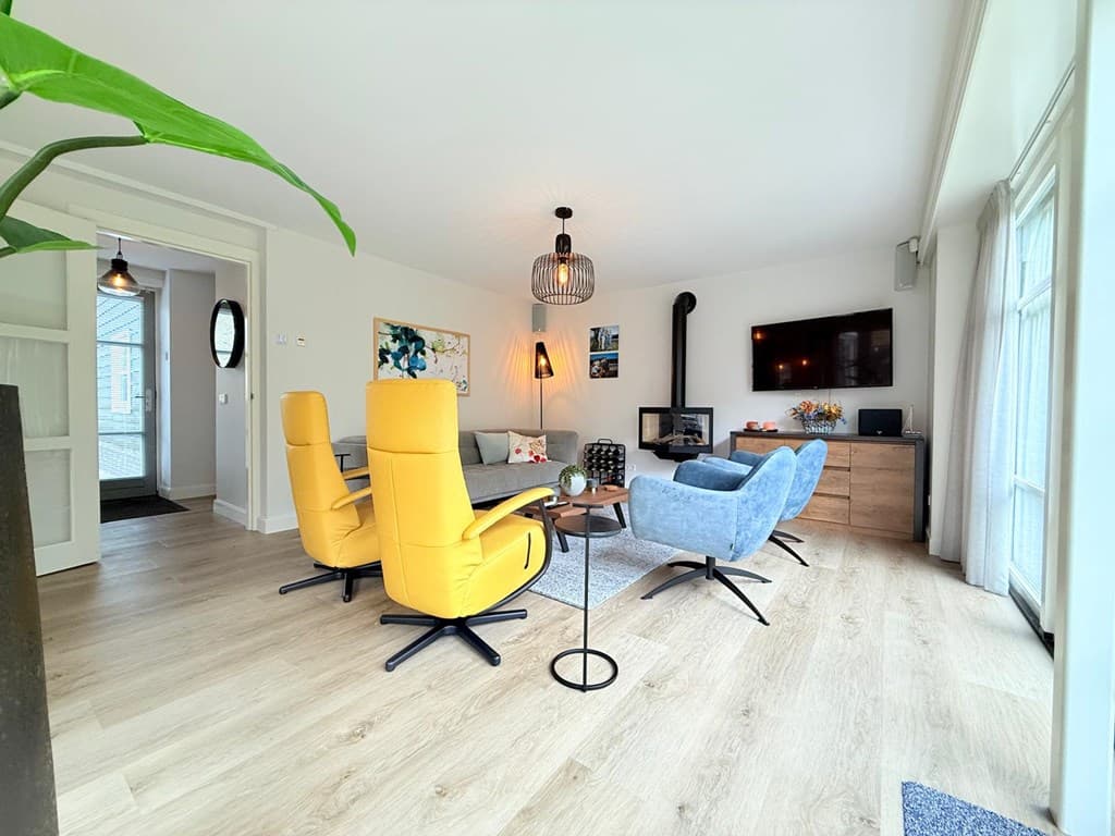 Moderne woonkamer met gele en blauwe fauteuils, een televisie, een kast en veel natuurlijk licht vanuit de ramen