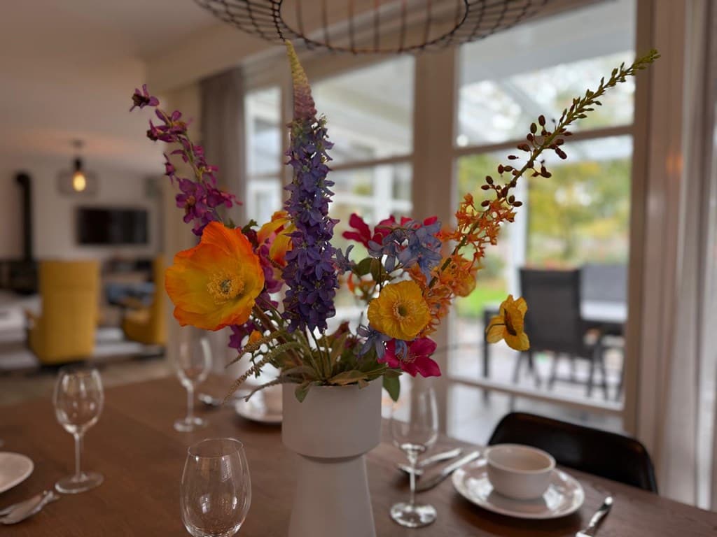 Kleurrijke bloemenarrangement in een vaas op een eettafel met glazen en servies, op de achtergrond een lichte woonkamer.