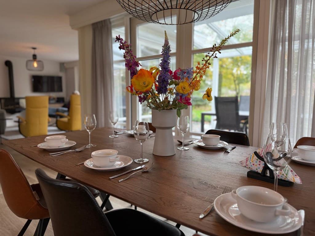 Eettafel met servies en een vaas met bloemen, zicht op een lichte woonkamer met banken en grote ramen