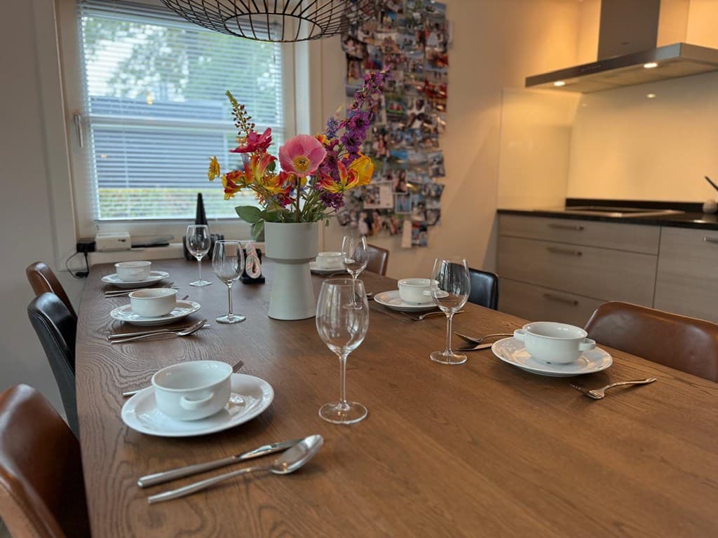 Eetkamer met een houten tafel, zes borden, glazen en een vaas met bloemen als centerpiece.