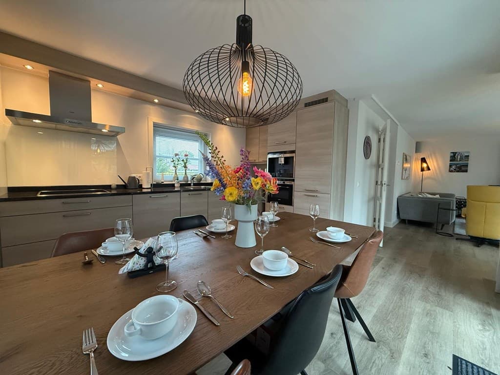 Ruime eetkamer met een houten tafel, moderne keuken in de achtergrond en een grote lamp boven de tafel met bloemenarrangement