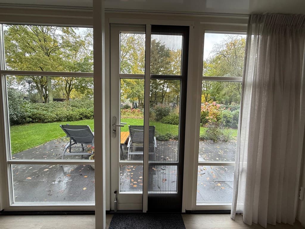 Zicht op een tuin met groene grasvelden en tuinmeubelen vanuit een woning met grote glazen deuren.