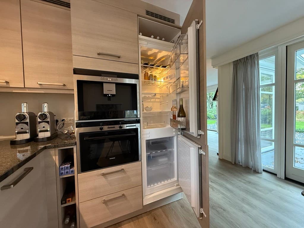 Keuken met een open koelkast en ingebouwde oven, lichte houttinten en een grote glazen deur naar buiten.