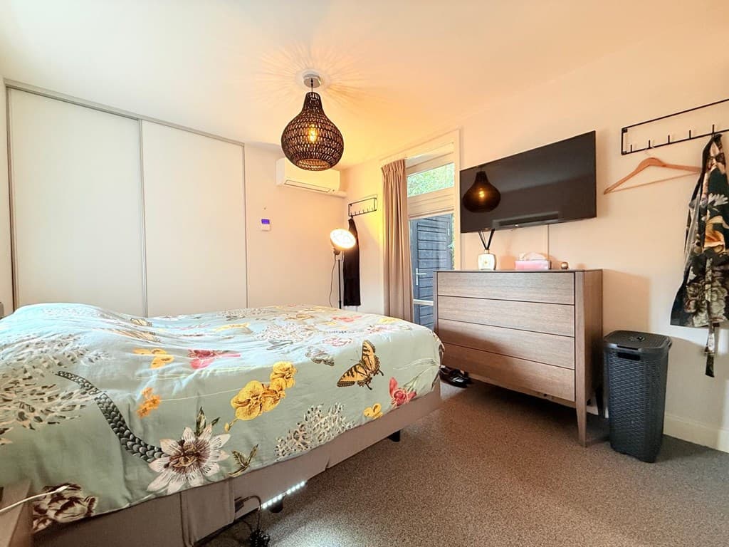 Slaapkamer met een tweepersoonsbed, kast, tv, lamp en stoel bij een voordeur naar buiten.