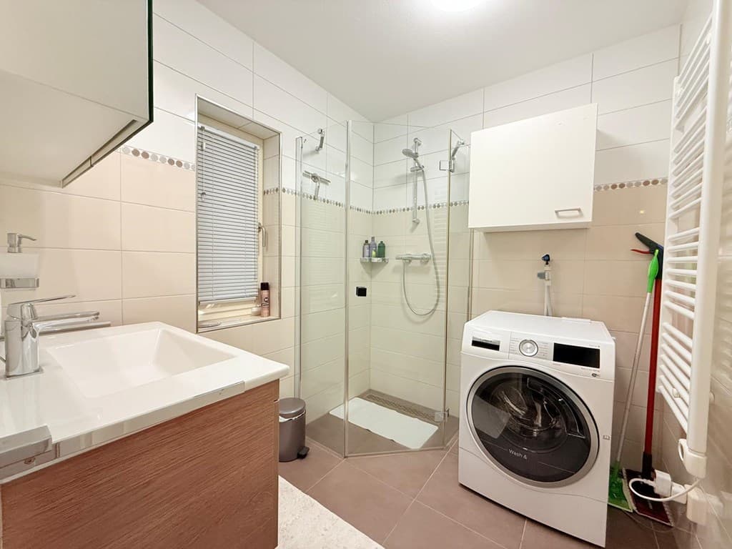 Moderne badkamer met douche, wastafel en wasmachine, afgewerkt met lichtetegels en een raam voor natuurlijke lichtinval