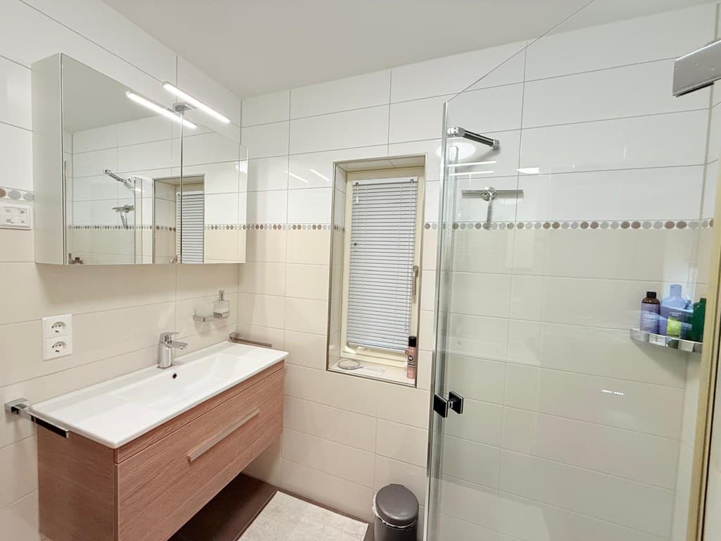 Moderne badkamer met inloopdouche, wastafel met spiegel en raam, en lichte tegelwand.