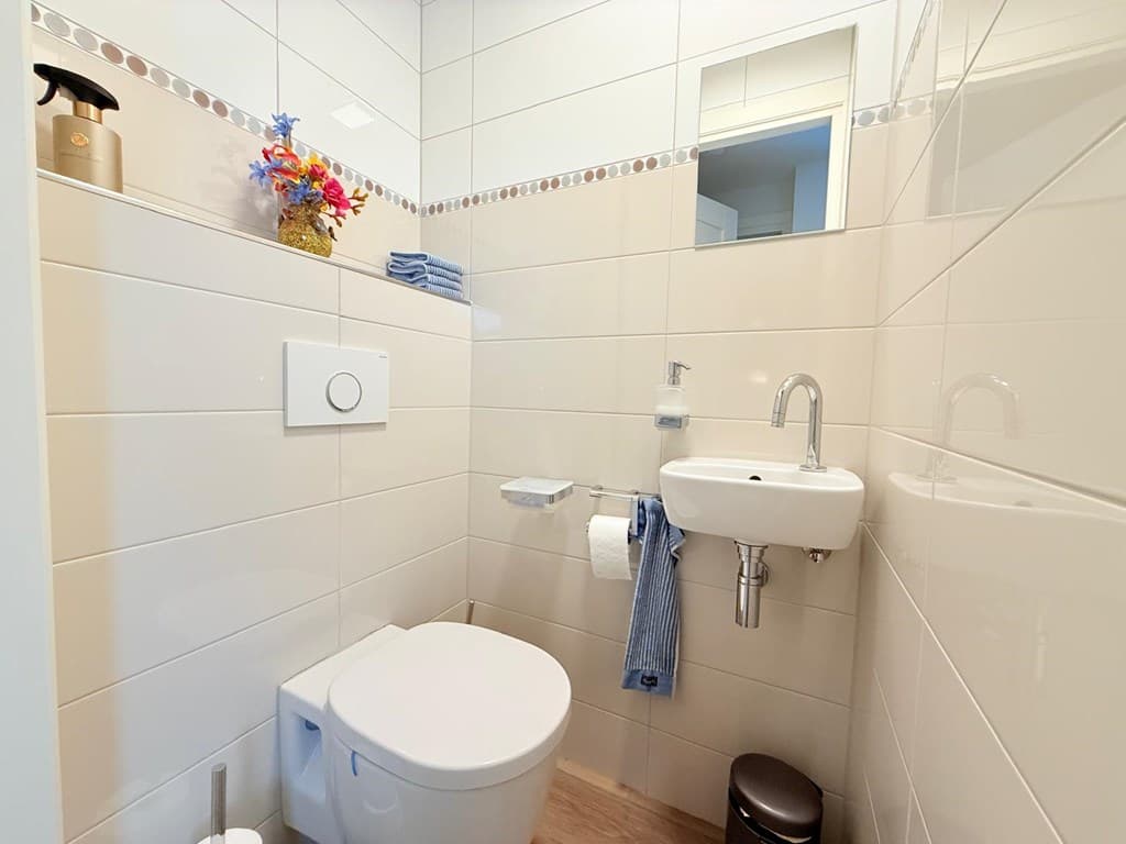 Badkamer met toilet, wastafel en spiegel, lichte wandtegels en decoratieve bloemen boven de wastafel.