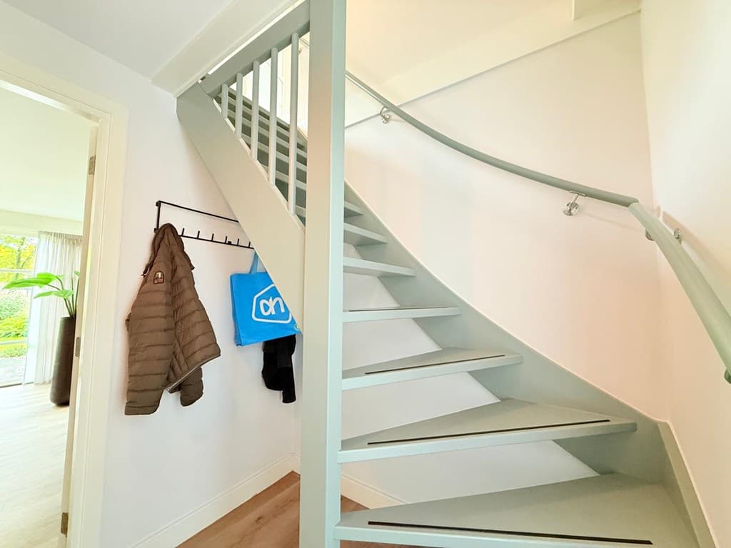 Trap met ronde leuning en kledinghangers in een lichte entree van de woning.
