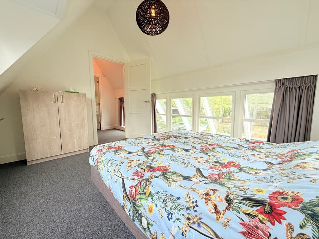 Slaapkamer met een groot bed met bloemenbedekking, kast en schuin dak met grote ramen die veel licht binnenlaten