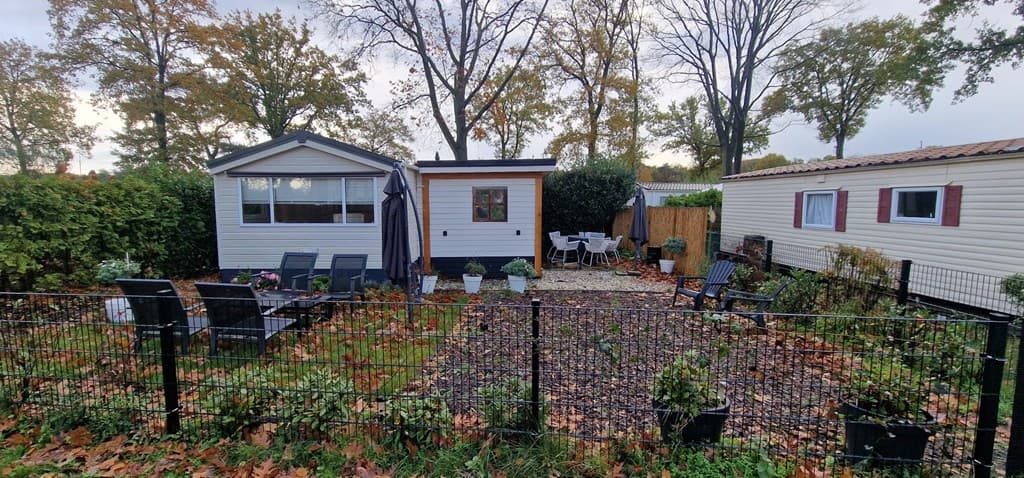 Tuin met terrasmeubilair, omheind grasgebied en twee vakantiehuizen, omringd door bomen en herfstbladeren.