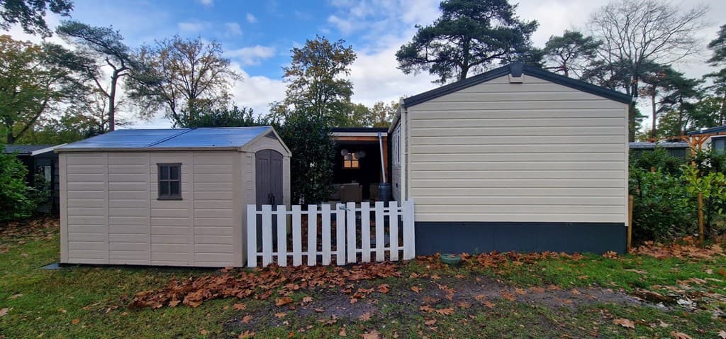 Twee chalets met lichtgekleurde houten wanden, een schuur en een hek, omringd door bomen en herfstbladeren.