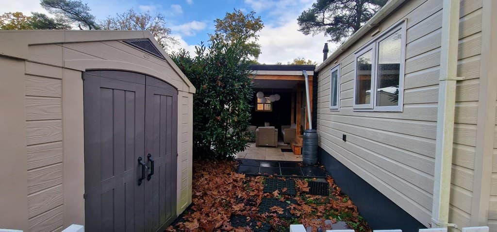 Zij-aanzicht van een vakantiewoning met een tuinhuisje en een terras met meubels, omgeven door bladeren en bomen.