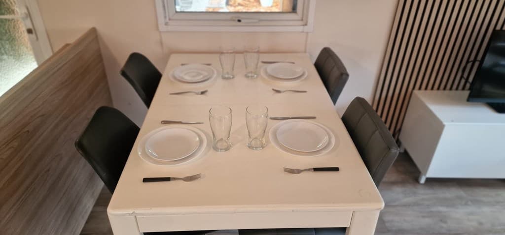 Eenvoudige eettafel met servies en glazen, omringd door zwarte stoelen en een tv-kast in de achtergrond