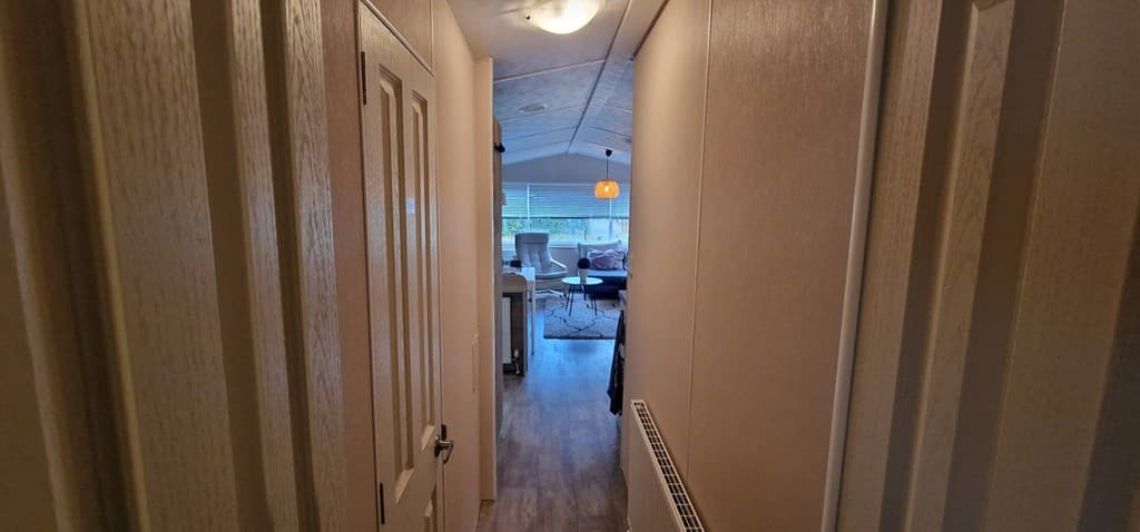 Zicht op een gang met een houten vloer en deuren, uitkomend in een lichte woonkamer met een zithoek en raampartij