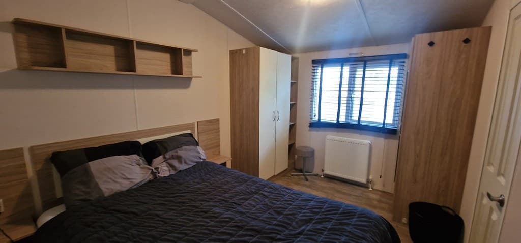 Slaapkamer met dubbelbed, houten kast en bureau, raam met jaloezieën en strakke vloer.