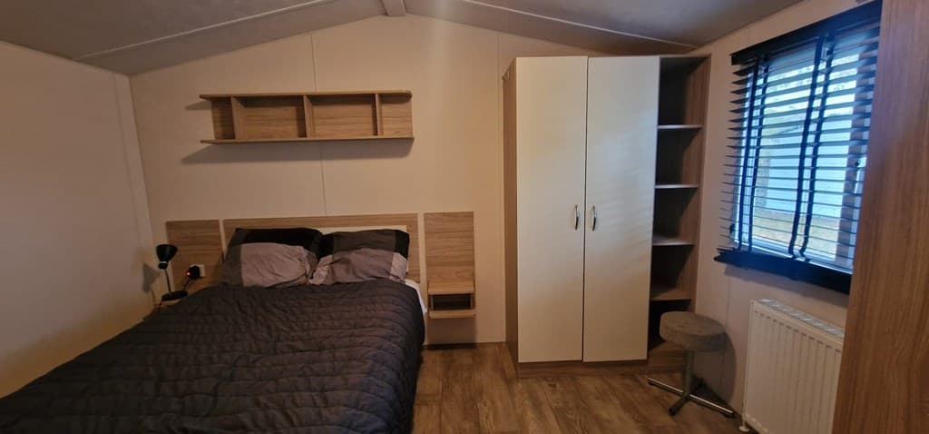Slaapkamer met een dubbele bed, kast en bureau, voorzien van een raam met jaloezieën en houten vloer.