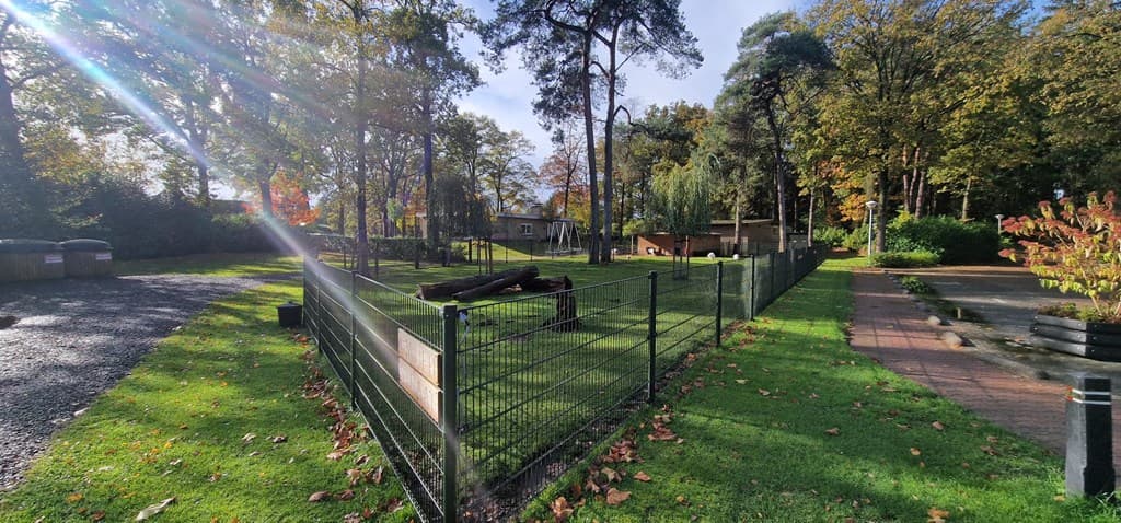 Groen grasveld omheind met een hek, bomen op de achtergrond en een eenvoudige woning zichtbaar tussen de natuur.