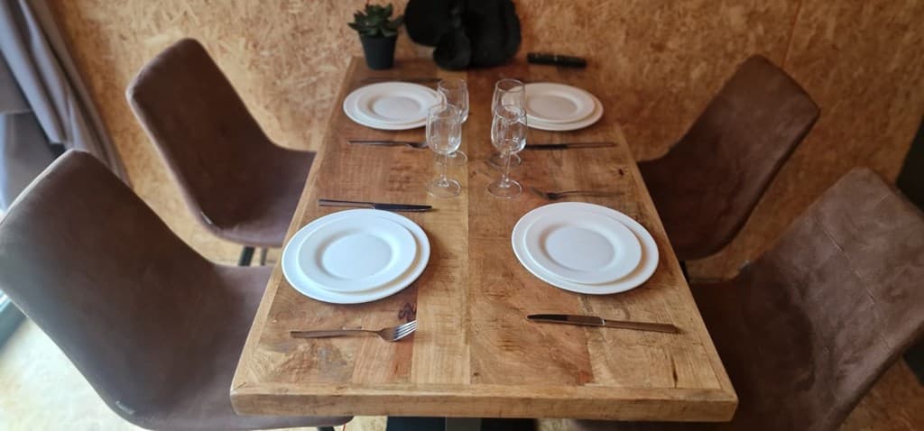 Eetkamertafel met vier stoelen, glazen en borden op een houten vloer met wand van geïmpregneerd hout.