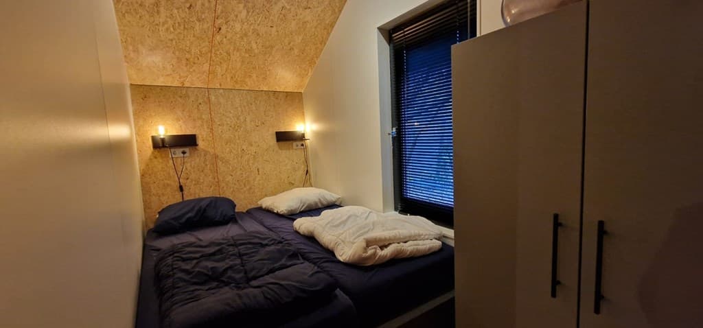 Slaapkamer met een tweepersoonsbed, nachtkastjes met verlichting en een raam met zwarte jaloezieën.