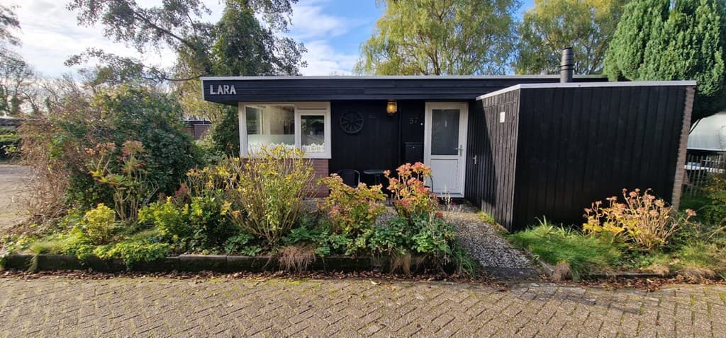Buitenzicht van een vakantiewoning met zwarte gevel, plantenbedden en een zithoek voor de deur.