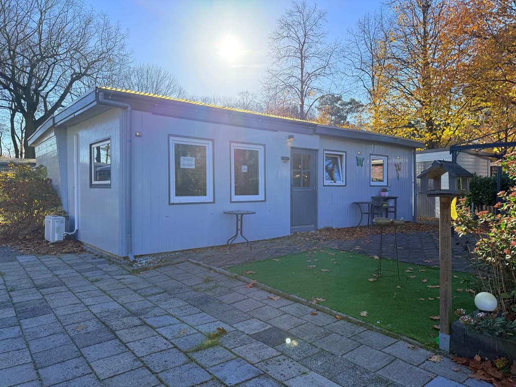 Zicht op een blauwe vakantiewoning met een betegelde oprit en rondom planten en bomen in het zonlicht.