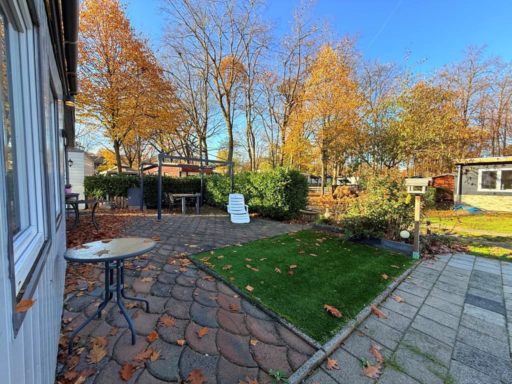 Tuin bij een woning met een terras, grasveld, bomen en herfstbladeren onder een blauwe lucht.