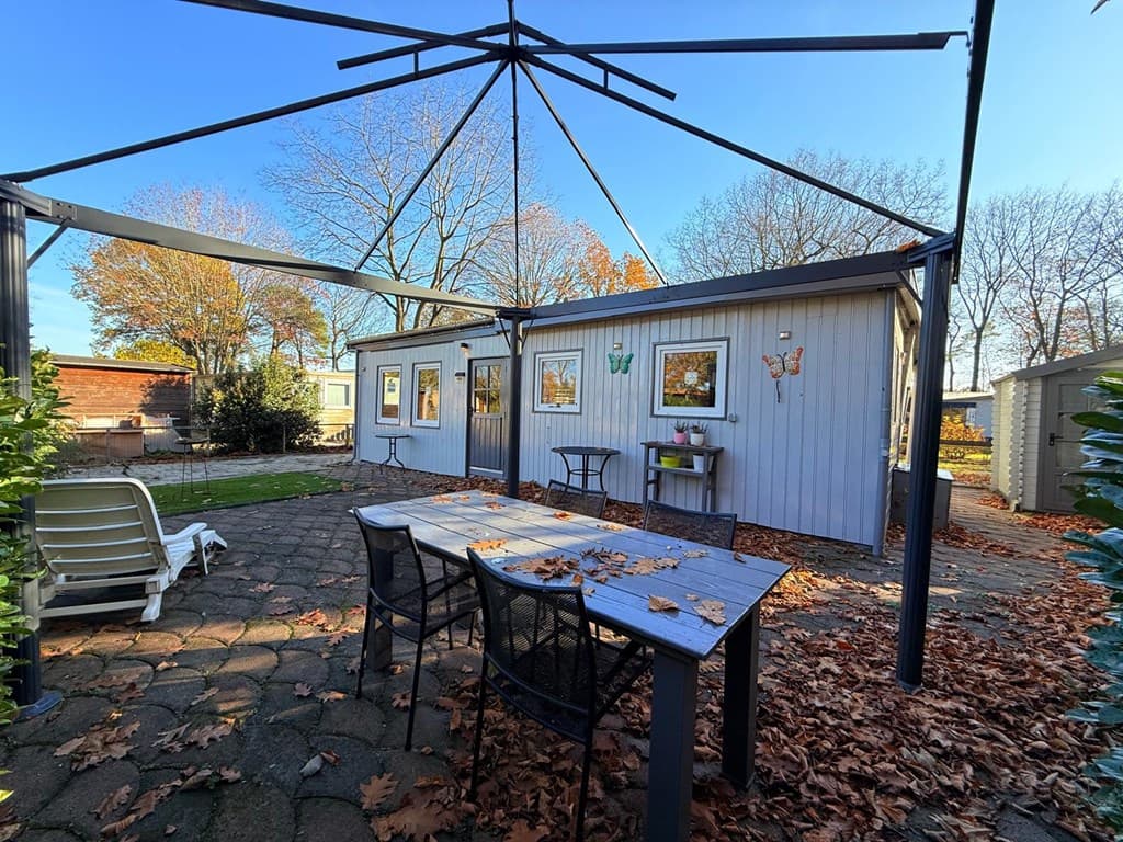 Tuintje met een betegeld terras, tuinset en een houten chalet in een groene omgeving met bomen en vallende bladeren