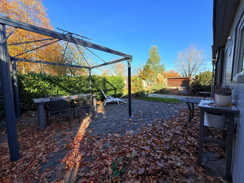 Terras met een overkapping, tuintafel en stoelen, omgeven door herfstbladeren en groene heggen.