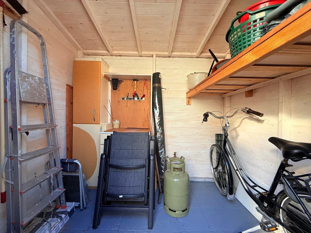 Binnenkant van een opbergschuur met een ladder, fietsen, een gasfles en enkele opbergkasten tegen de wand