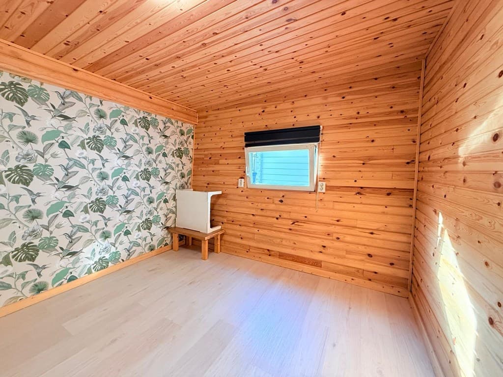 Houten ruimte met een behang van groene bladeren, een raam en een houten bankje tegen de muur.