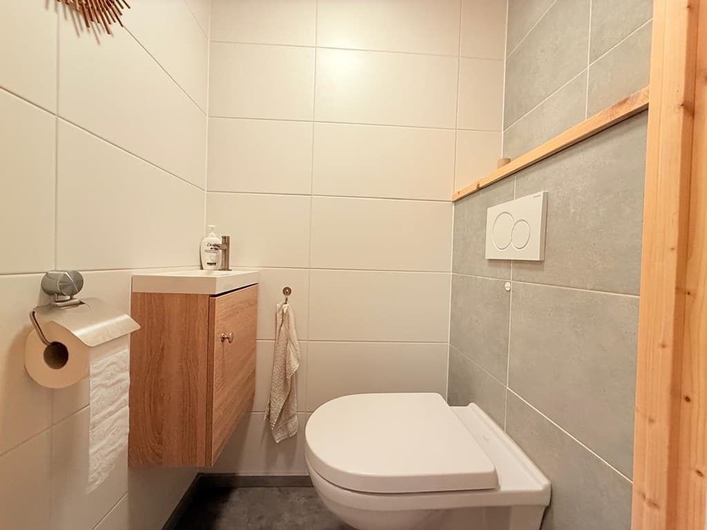 Toilet met wand- en vloerbedekking in neutrale kleuren, een houten opbergmeubel en handdoekhaakje aan de muur