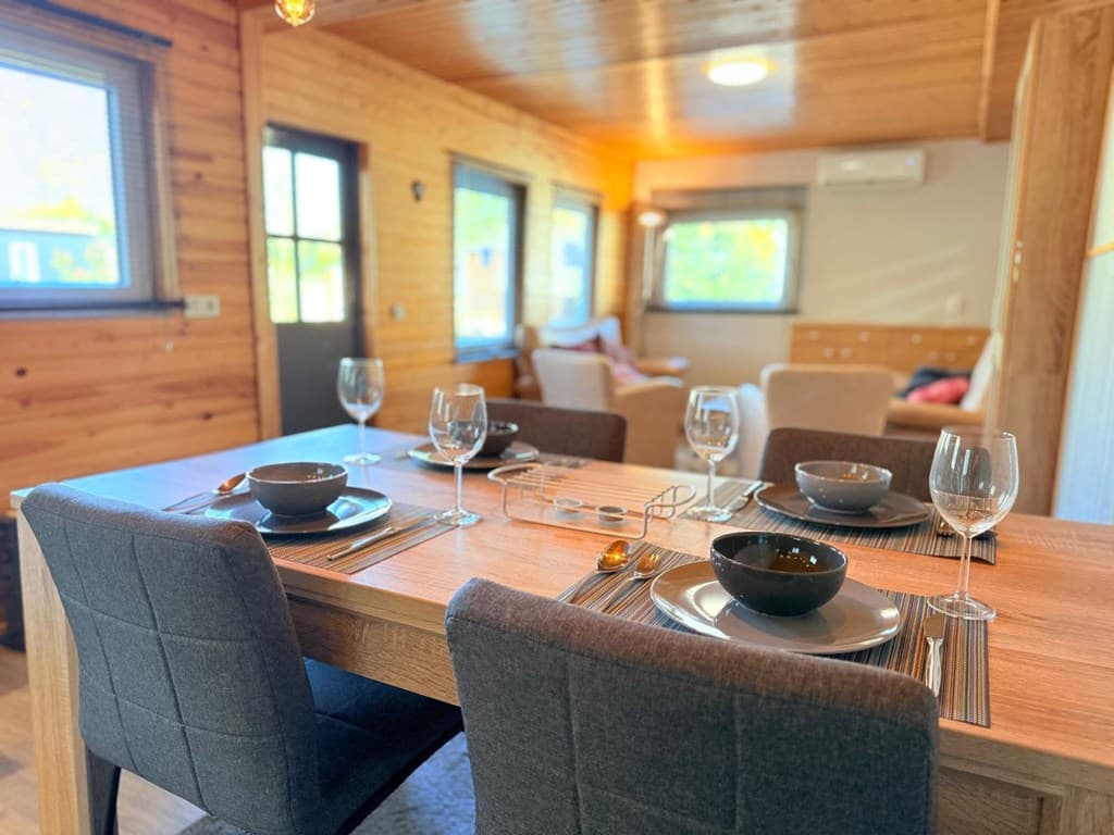 Eethoek met een houten tafel, glazen en borden, en een uitzicht op een lichte woonkamer met houten panelen