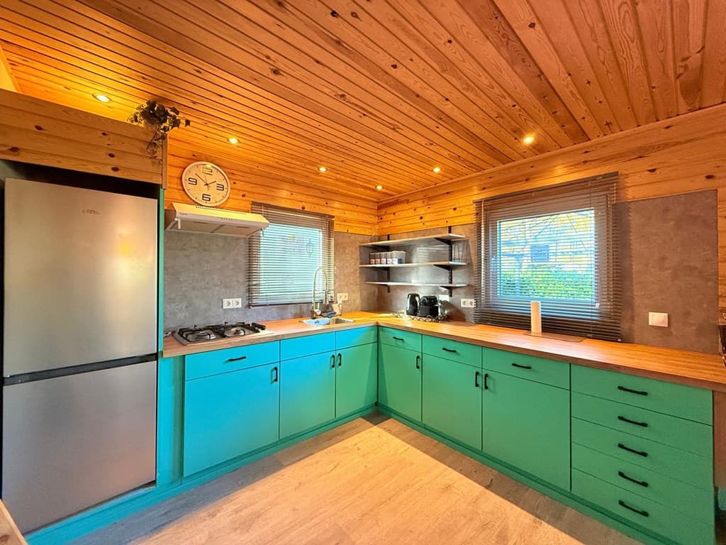 Moderne keuken met houten plafond, groene kasten, kookplaat en koelkast nabij een raam met uitzicht naar buiten