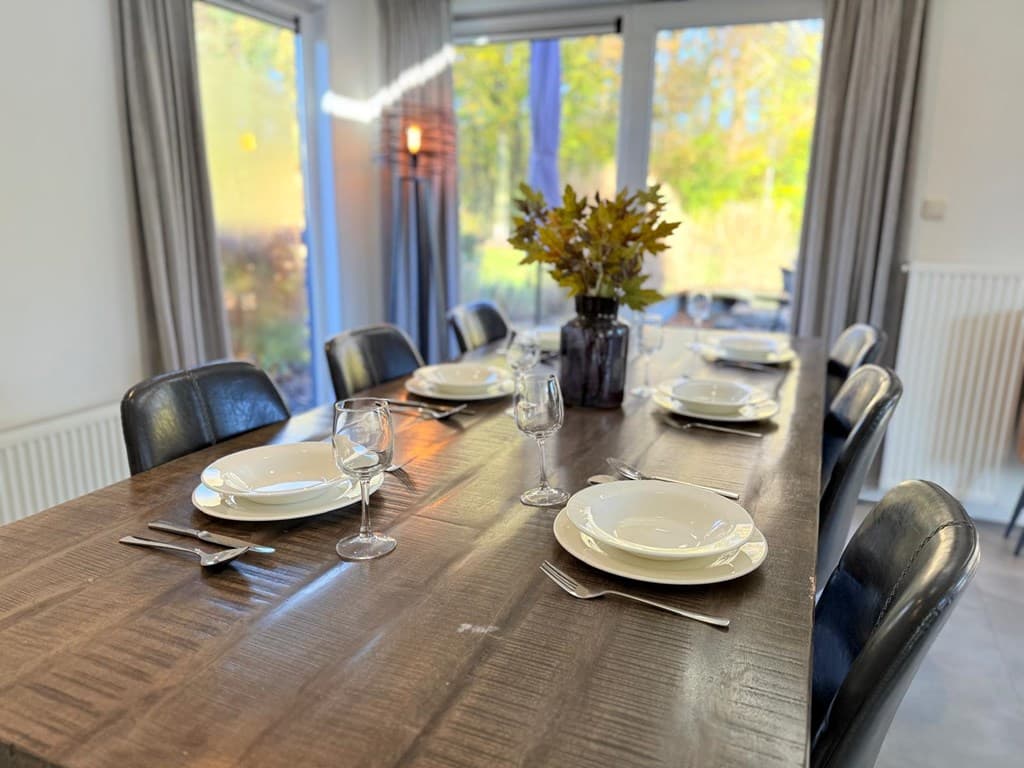 Eettafel met servies en glazen, uitzicht op een tuin door grote ramen met gordijnen aan beide zijden