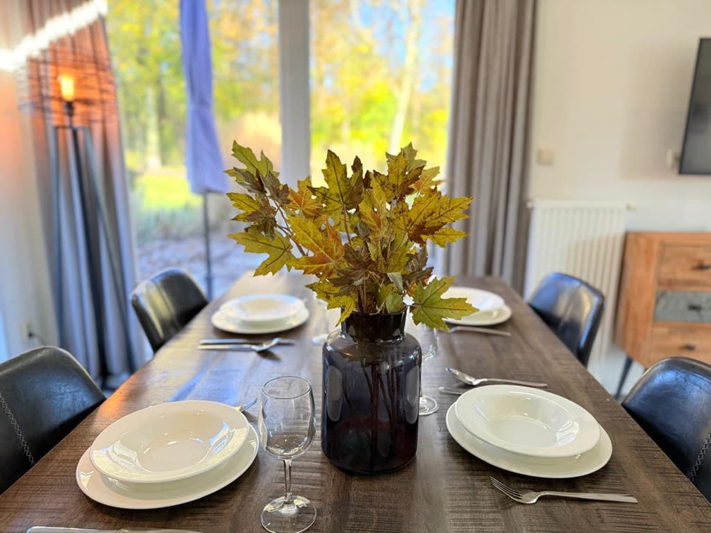 Eettafel met borden en glazen, een vaas met gele bladeren staat in het midden en grote ramen geven uitzicht op een groene omg