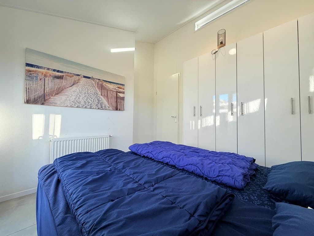 Lichte slaapkamer met een tweepersoonsbed, witte kast en een foto van een strand op de muur.
