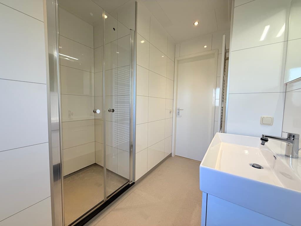 Moderne badkamer met glazen douchecabine, wastafel en lichte wand- en vloertegels.