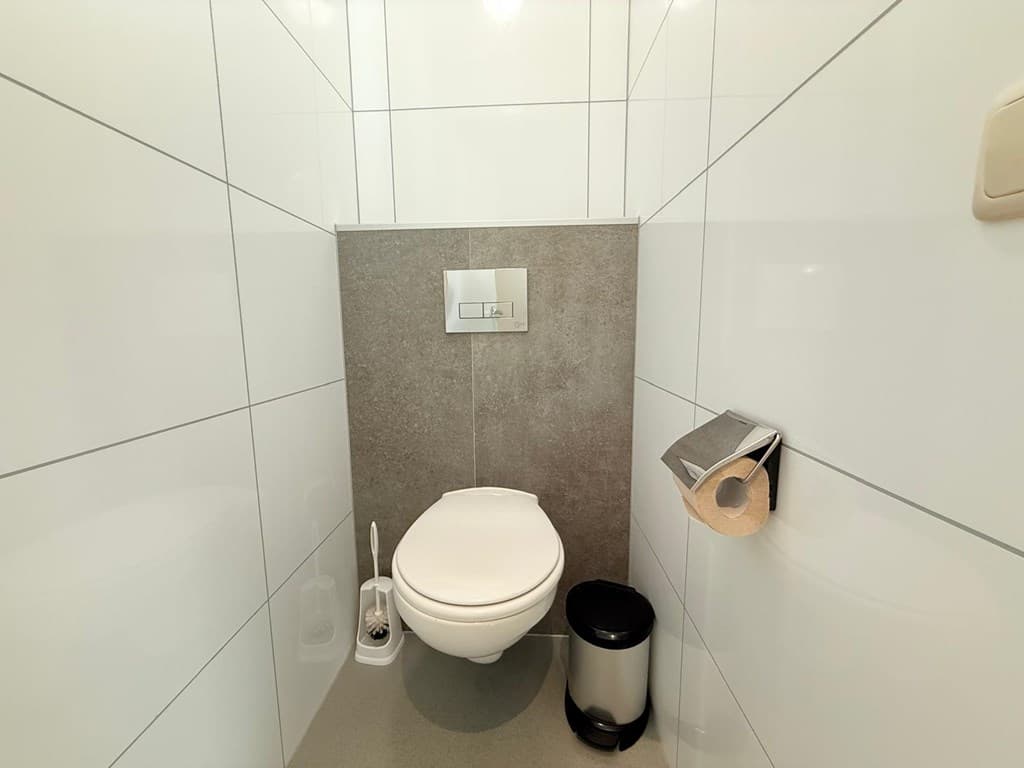 Modern toilet met witte tegels, grijze wand en afvalbak, voorzien van toiletpapierhouder en toiletborstel.
