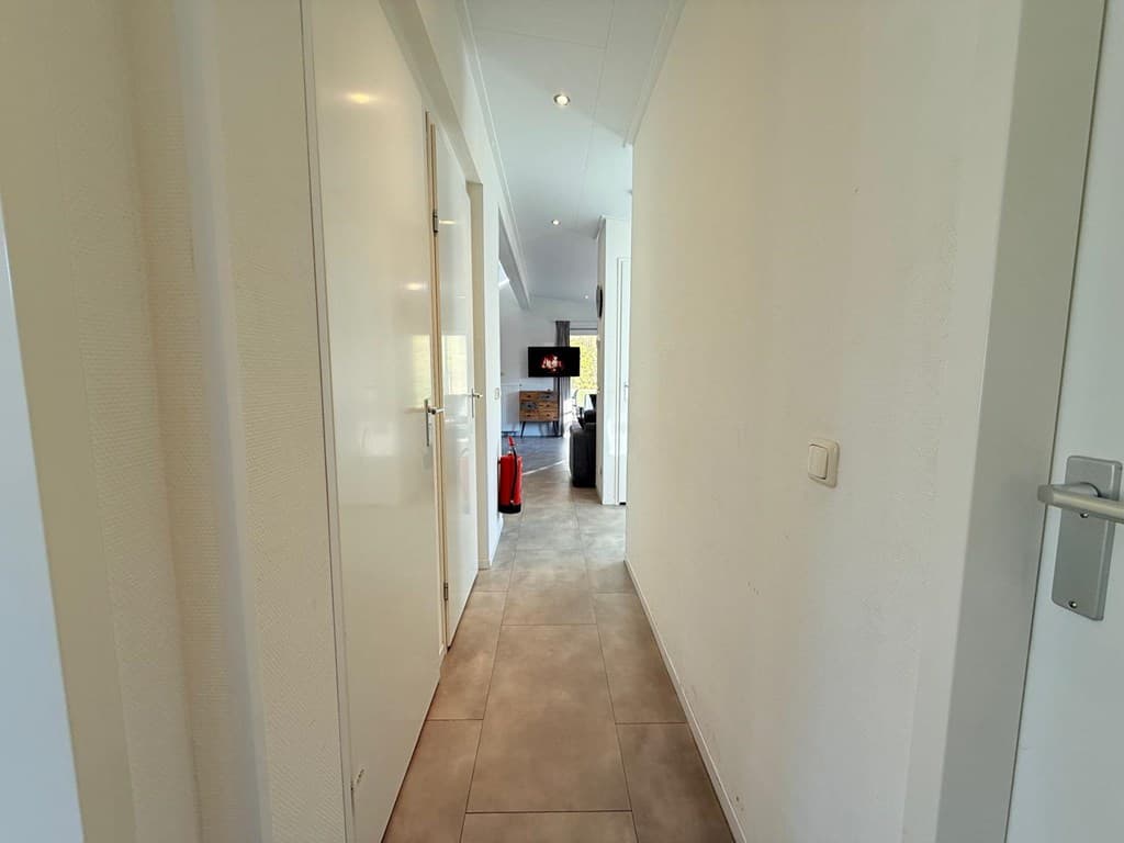 Een smalle gang met witte wanden en tegelvloer, zicht op een open woonkamer met een televisie aan de eindzijde.