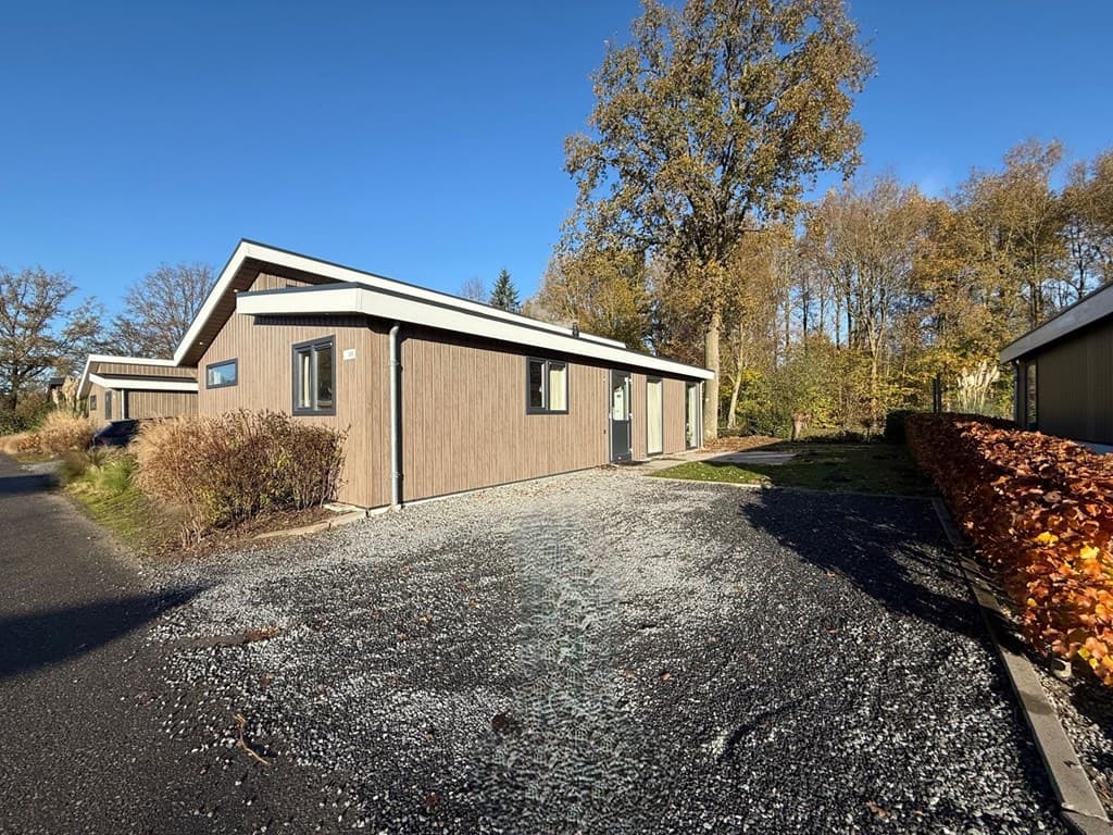 Moderne vakantiewoning met houten gevel, gelegen aan een verhard pad omringd door bomen en groen.