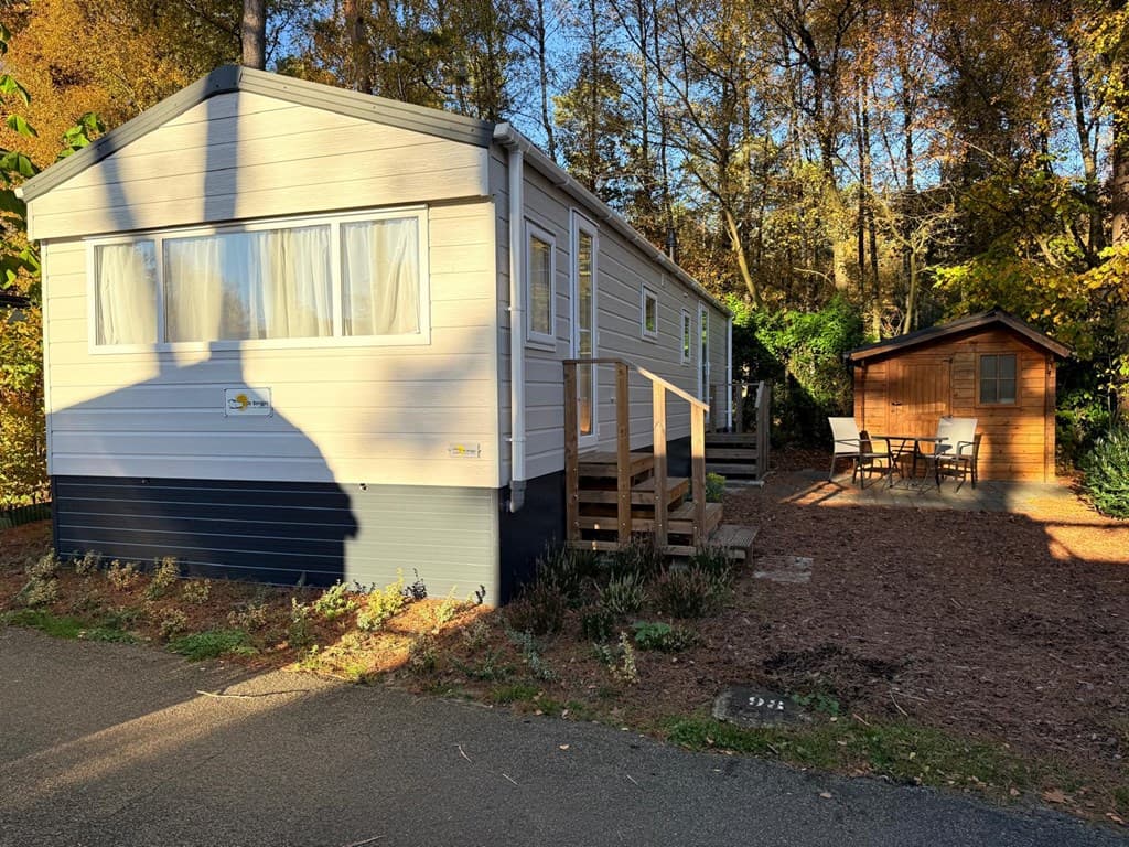 Moderne vakantiewoning met houten terras en een houten tuinhuisje, omringd door bomen en een grindpad.