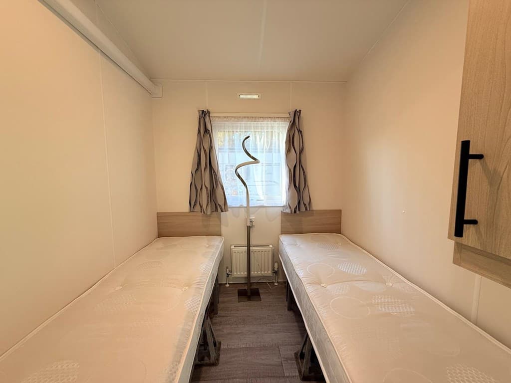 Slaapkamer met twee eenpersoonsbedden, een raam en een gordijn in lichte kleuren.