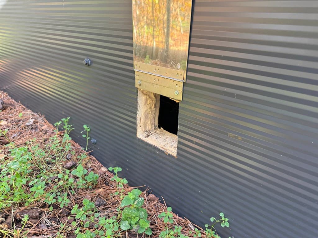 Opening in de zwarte wand van een woning met een metalen afwerking, omringd door groen en bomen