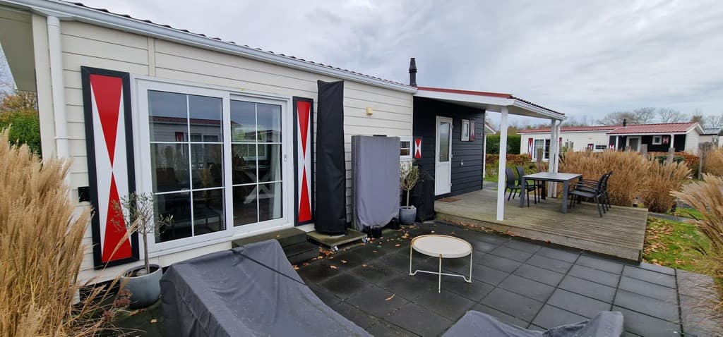 Buitenaanzicht van een vakantiehuis met een houten terras, grote ramen en tuinmeubilair, omringd door planten en gras.